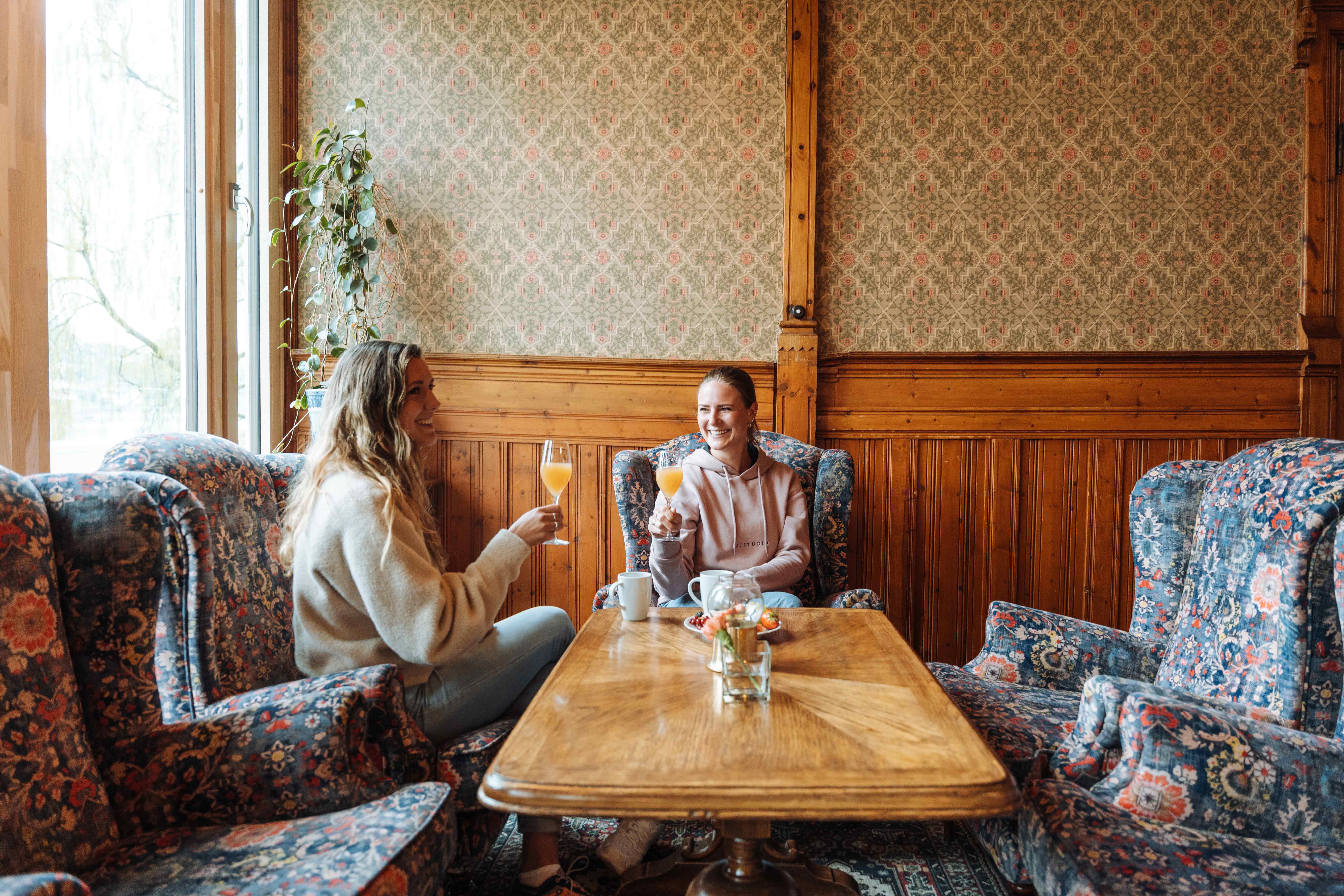 Two friends enjoy a glass of fresh juice in a charming lounge at Thon Hotel Sandven, surrounded by traditional Norwegian decor.