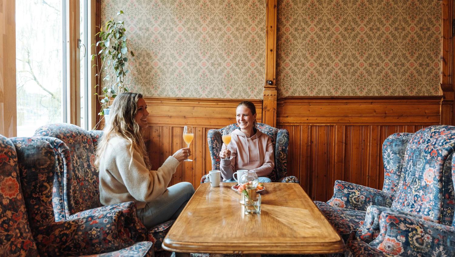 Two friends enjoy a glass of fresh juice in a charming lounge at Thon Hotel Sandven, surrounded by traditional Norwegian decor.