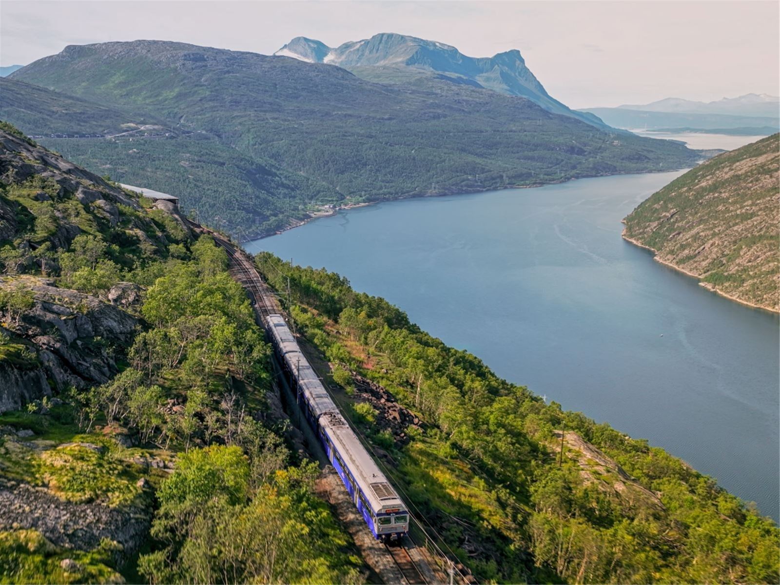 A passenger train runs along tracks on a rocky hillside beside a blue fjord, with green mountains in the distance.