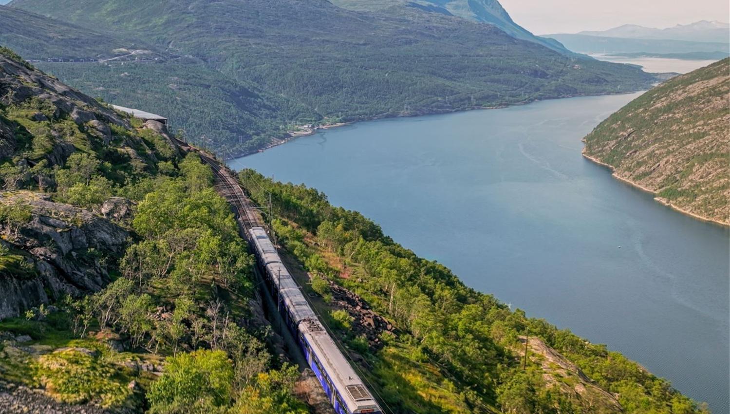 A passenger train runs along tracks on a rocky hillside beside a blue fjord, with green mountains in the distance.