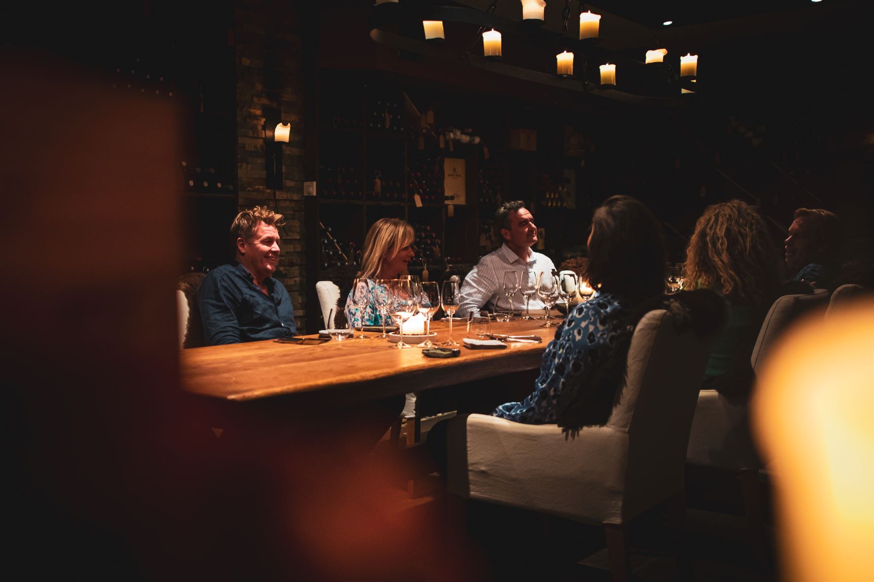 A group of persons sitting at a long table with wine glasses on it and a wall with wine bottles on shelves in the background