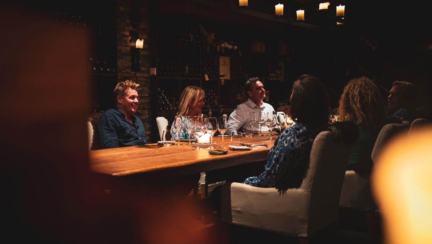 A group of persons sitting at a long table with wine glasses on it and a wall with wine bottles on shelves in the background