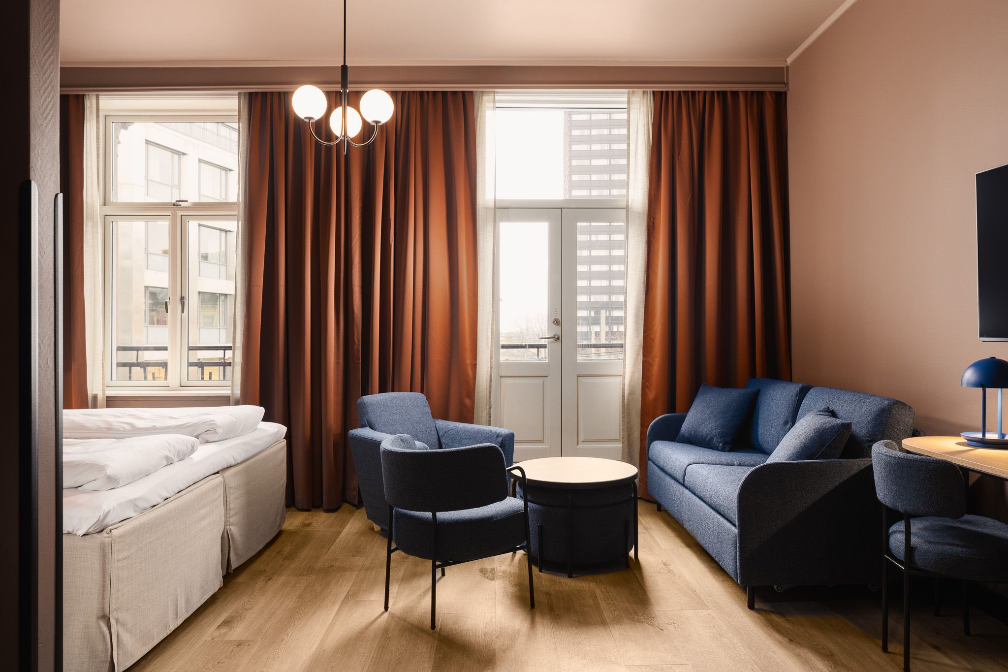 An interior of a family room with a large double bed, a blue sofa, blue armchairs, a table, brown curtains, and a balcony
