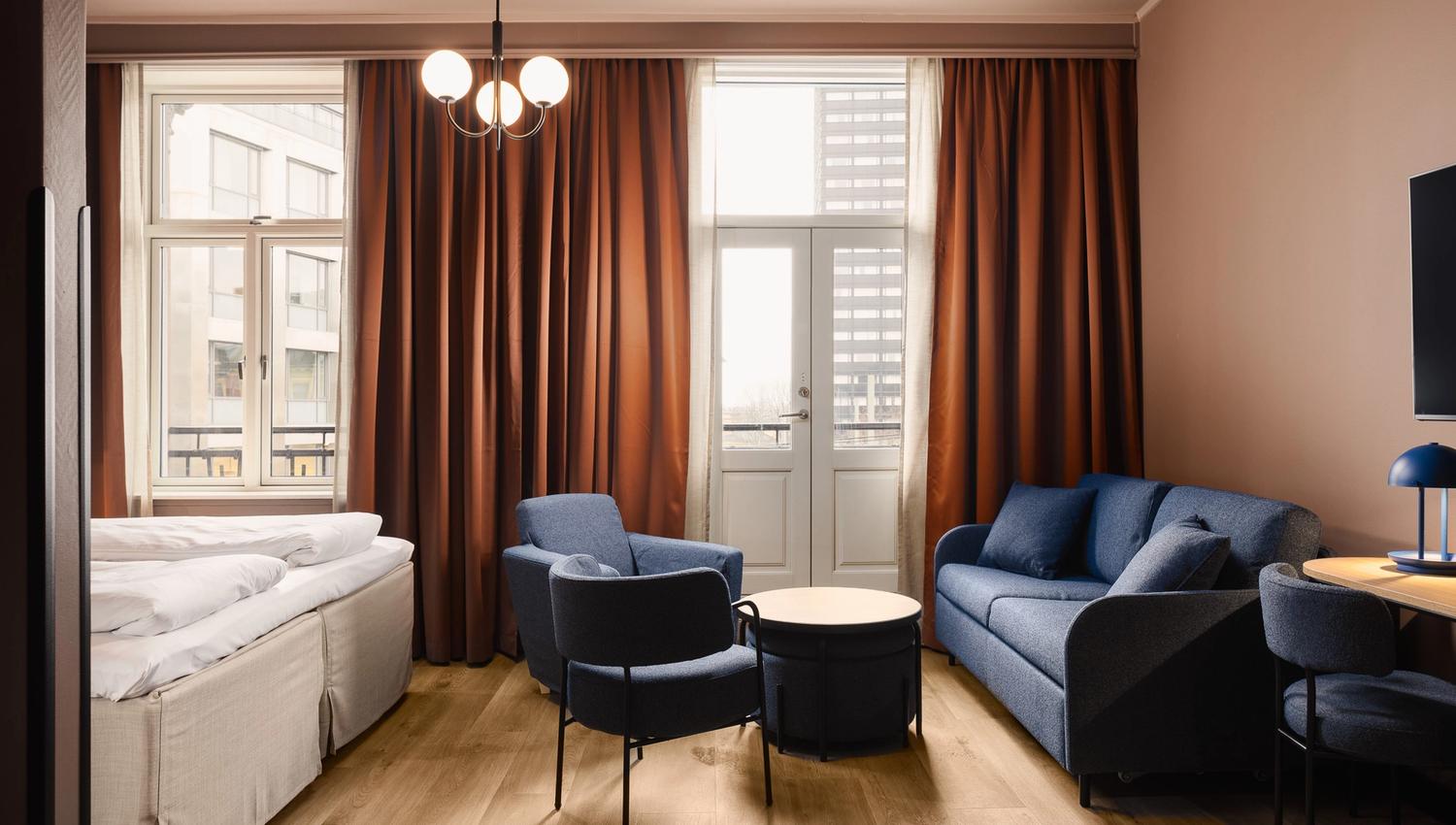 An interior of a family room with a large double bed, a blue sofa, blue armchairs, a table, brown curtains, and a balcony