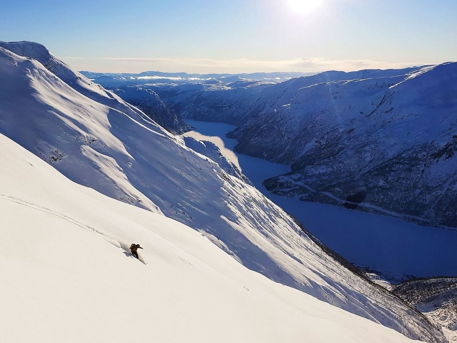 Een eenzame skiër snijdt een steile, besneeuwde helling af boven een kronkelend bergmeer onder een heldere blauwe lucht en zon..