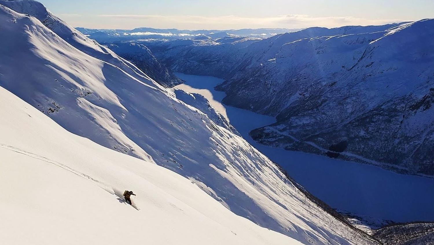 Een eenzame skiër snijdt een steile, besneeuwde helling af boven een kronkelend bergmeer onder een heldere blauwe lucht en zon..