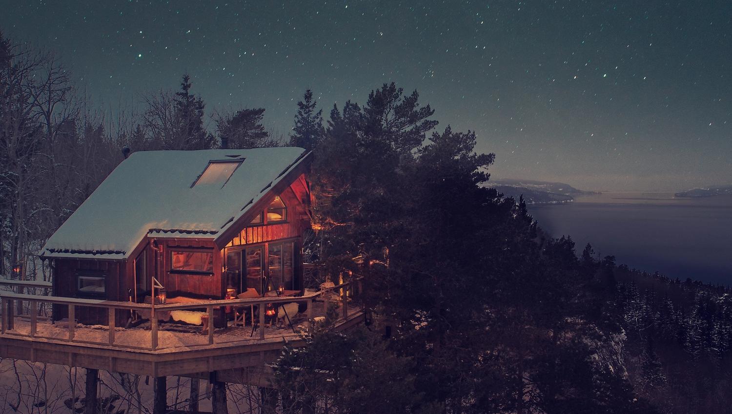 Drone photo with the Oslofjord in the background and an illuminated treetop cabin with many trees around.