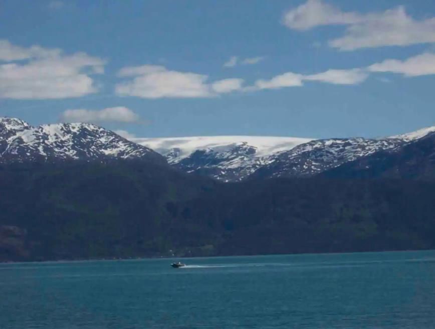 View over the Hardangerfjord with majestic snow-covered mountains and a boat on the crystal-clear water. In the background, the Folgefonna glacier, No