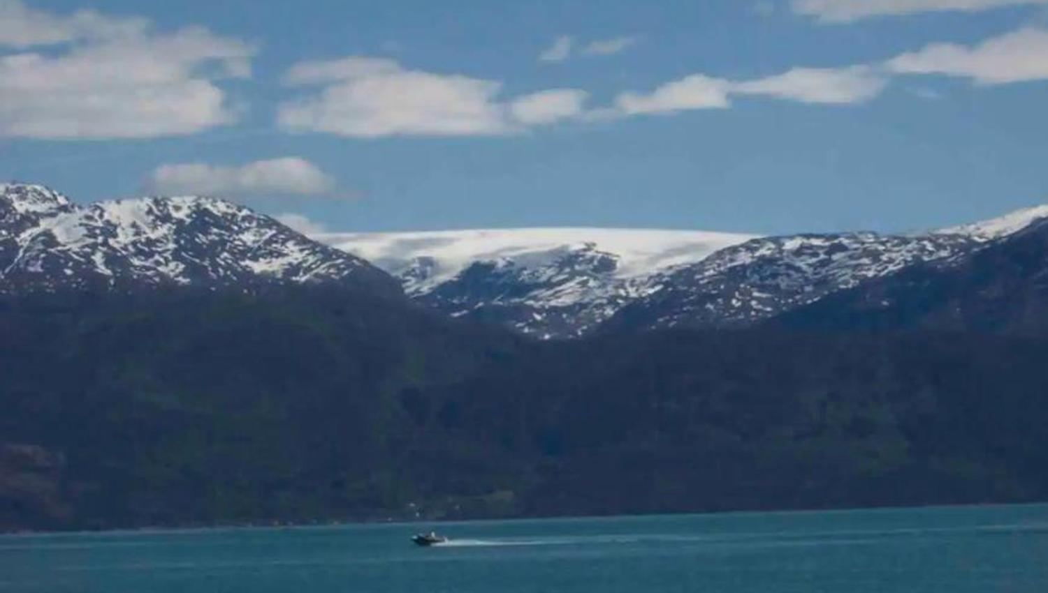 View over the Hardangerfjord with majestic snow-covered mountains and a boat on the crystal-clear water. In the background, the Folgefonna glacier, No
