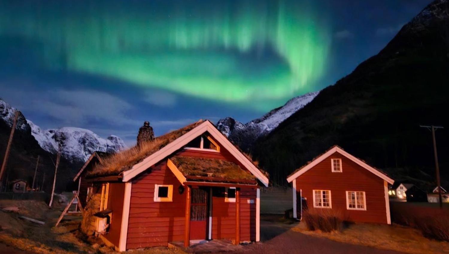 Green aurora swirls across a night sky above red wooden cabins, with snow-dusted mountains in the background at Aabrekk Gard Trollbu in Oldedalen valley..