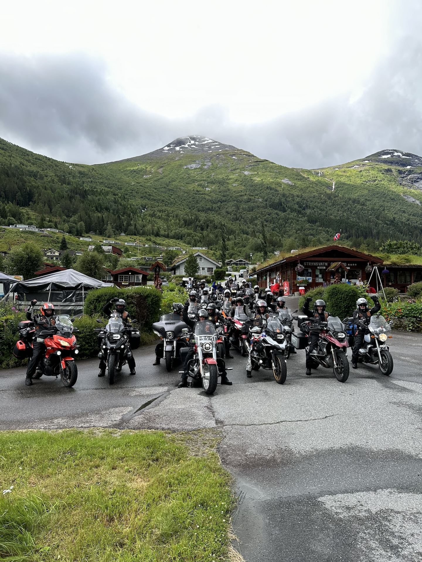Large group of motorcyclists stopped on a wet road in a village, with green mountains and cloudy sky behind..