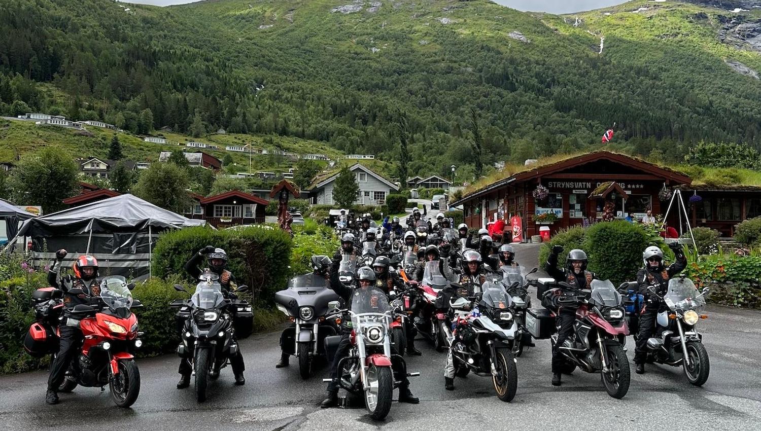 Large group of motorcyclists stopped on a wet road in a village, with green mountains and cloudy sky behind..