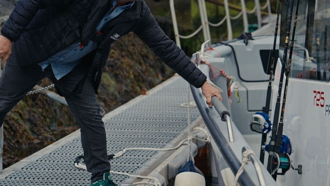 Person in a dark jacket steps onto a docked boat, holding the railing near fishing rods and coiled ropes..