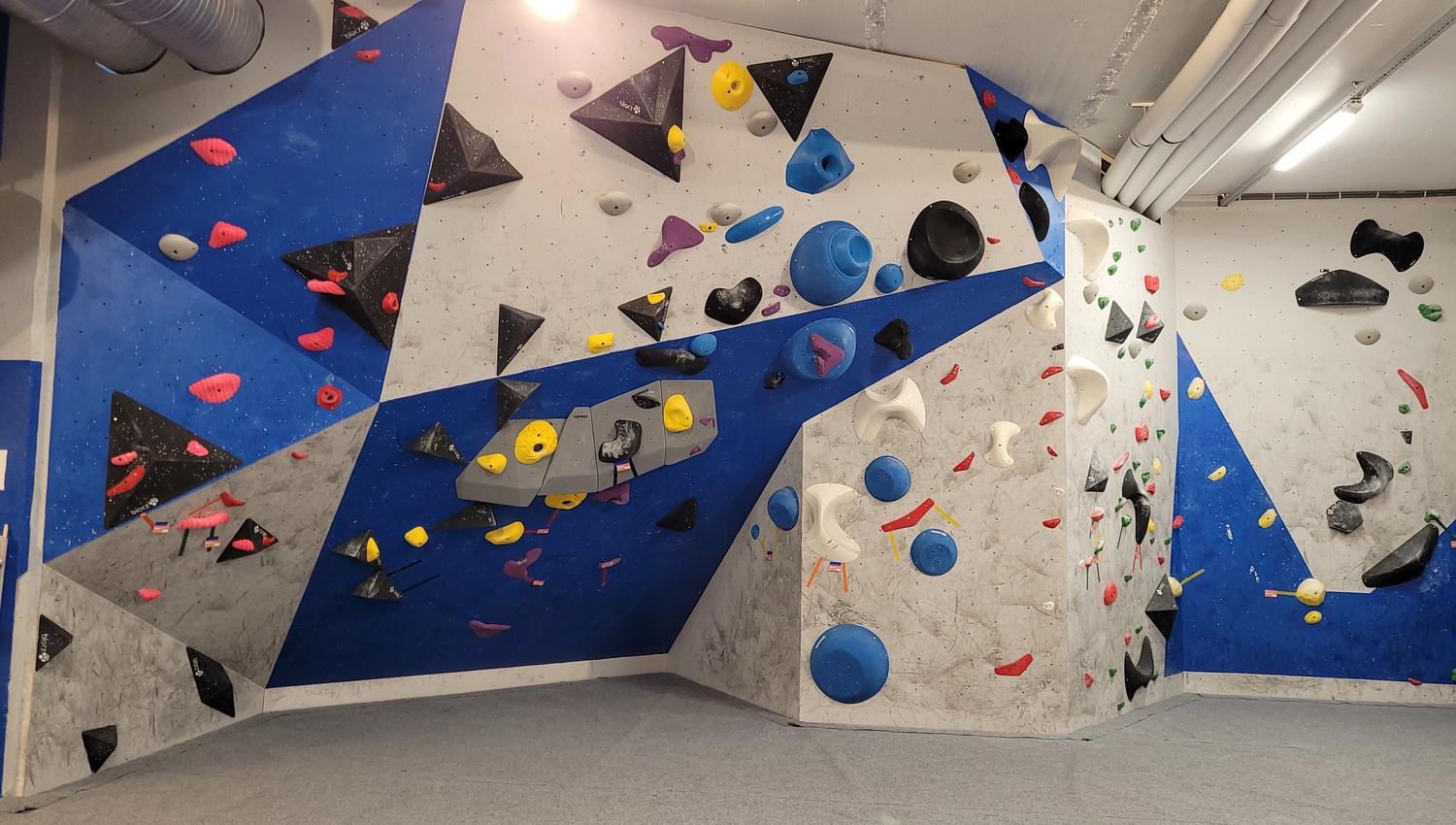 Indoor bouldering wall with blue and gray panels, assorted climbing holds and volumes, and padded floor below..