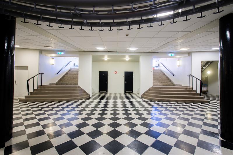 A large entrance hall with a black-and-white checkered floor and two staircases rising on each side. The walls are light, and the lighting is modern a