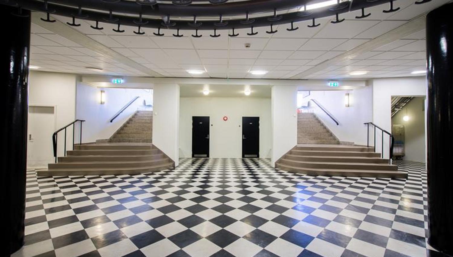 A large entrance hall with a black-and-white checkered floor and two staircases rising on each side. The walls are light, and the lighting is modern a