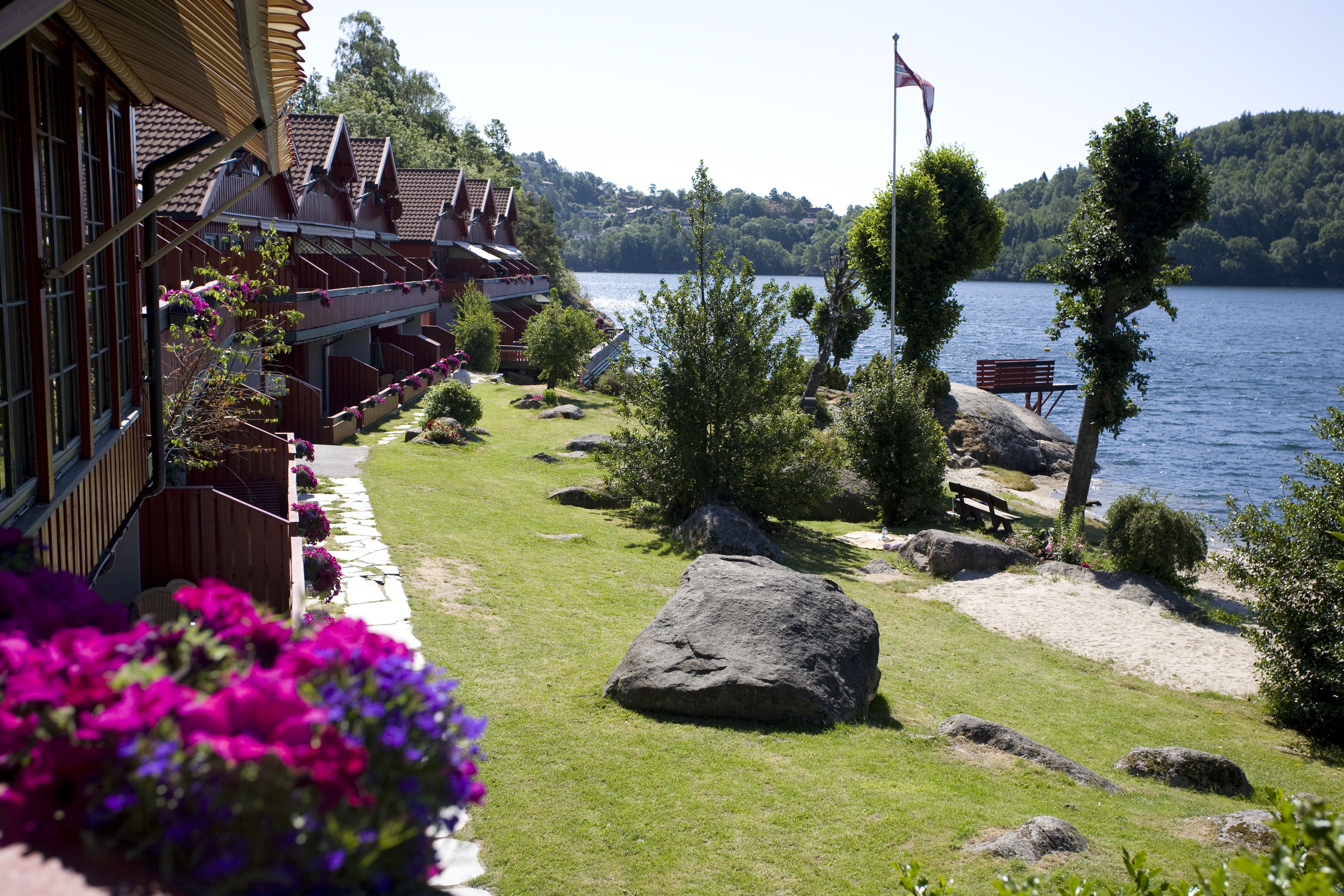 Holiday homes with pink-purple flowers in flower boxes on every terrace on the left side, freshly cut lawn, and water on the right. A Norwegian pennan