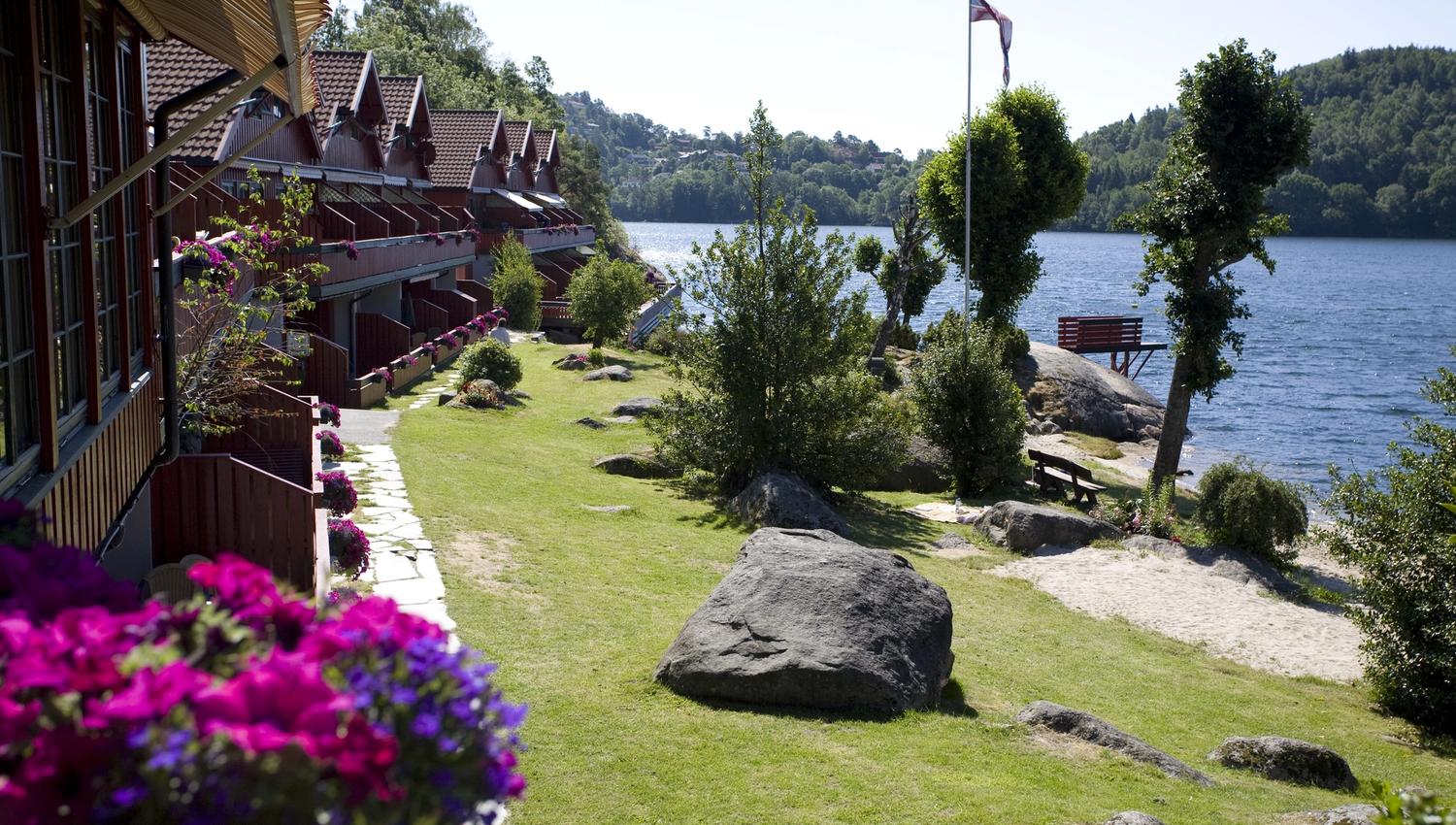 Holiday homes with pink-purple flowers in flower boxes on every terrace on the left side, freshly cut lawn, and water on the right. A Norwegian pennan
