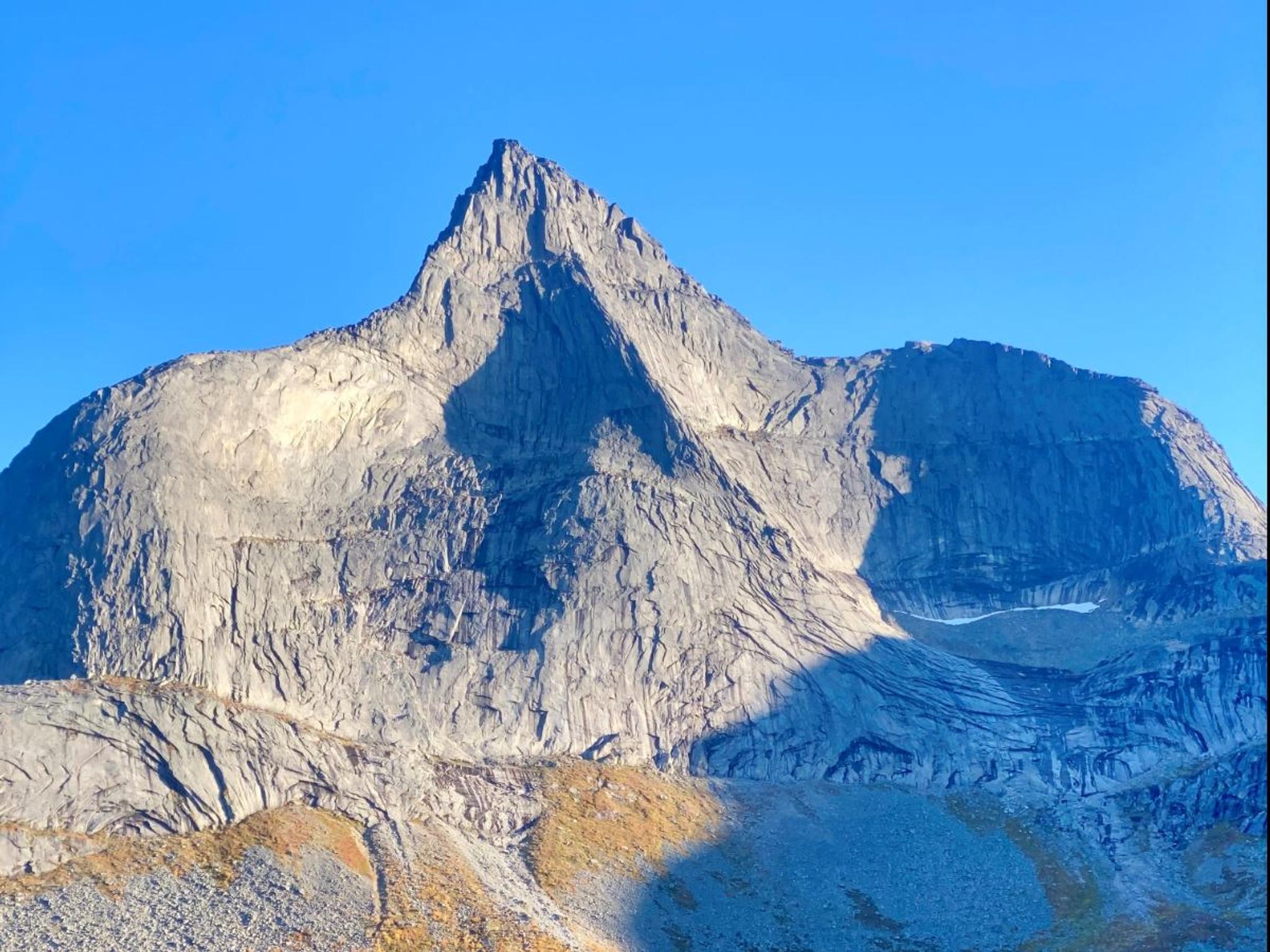 Close-up of a steep, grey mountain peak with shadows and stone scree in the foreground under a clear blue sky.