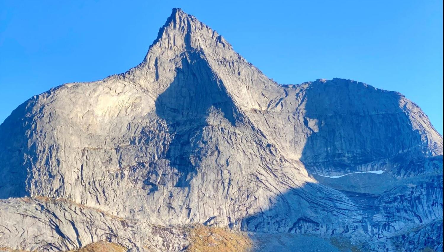 Close-up of a steep, grey mountain peak with shadows and stone scree in the foreground under a clear blue sky.