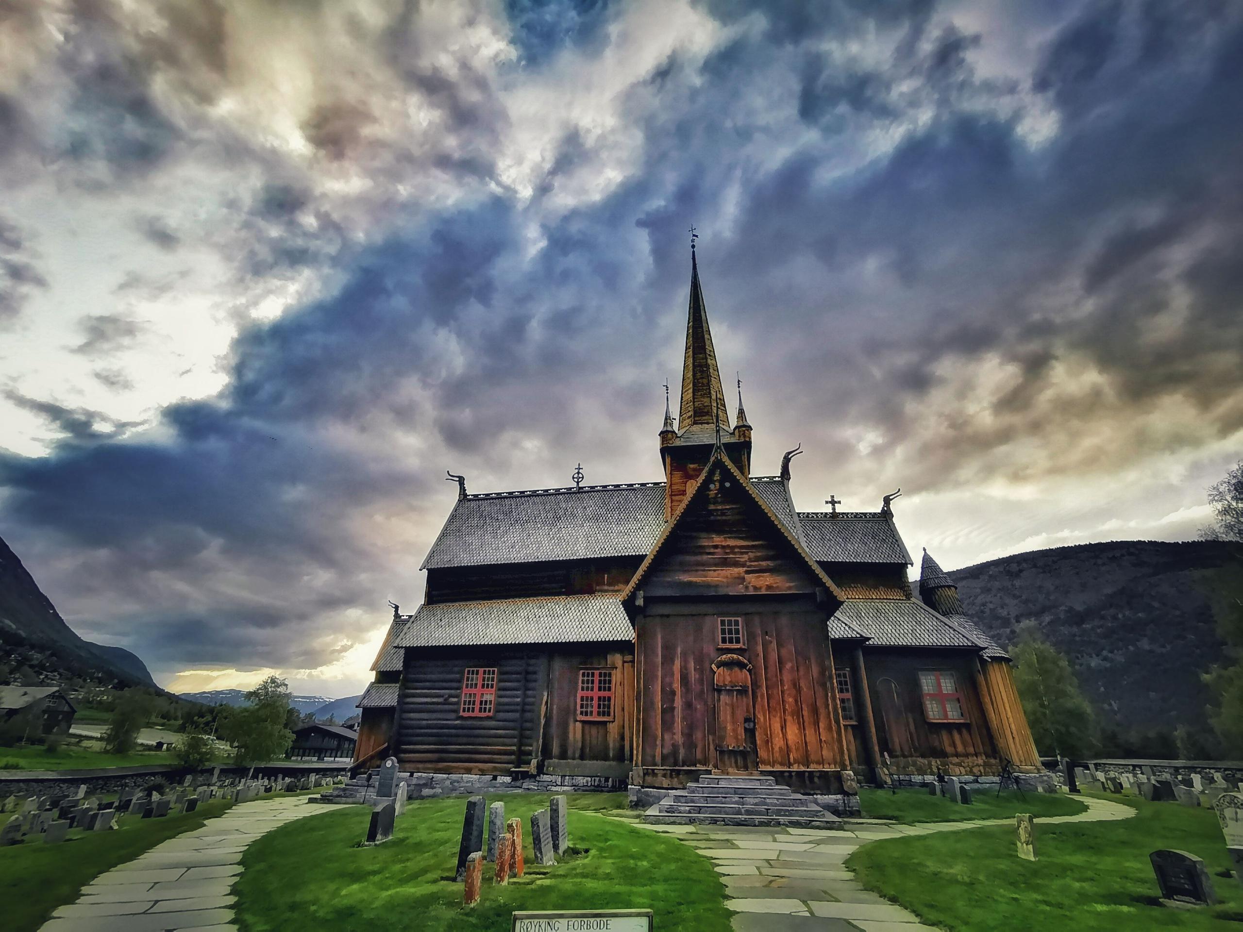 A dark wooden church with a tall spire stands amid a graveyard, with mountains and dramatic clouds overhead..