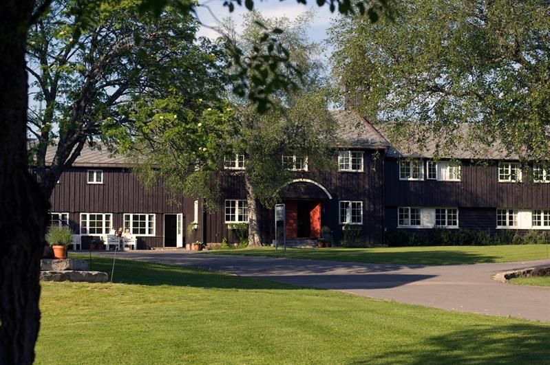 Lyseby facade. Brown wooden two-floor building with an arched doorway. Green lawn and trees in front. Photo