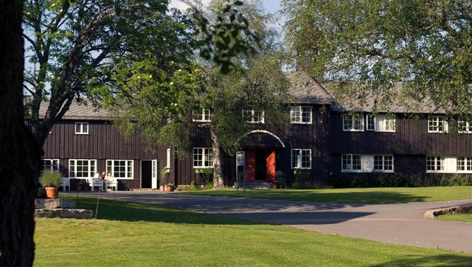 Lyseby facade. Brown wooden two-floor building with an arched doorway. Green lawn and trees in front. Photo