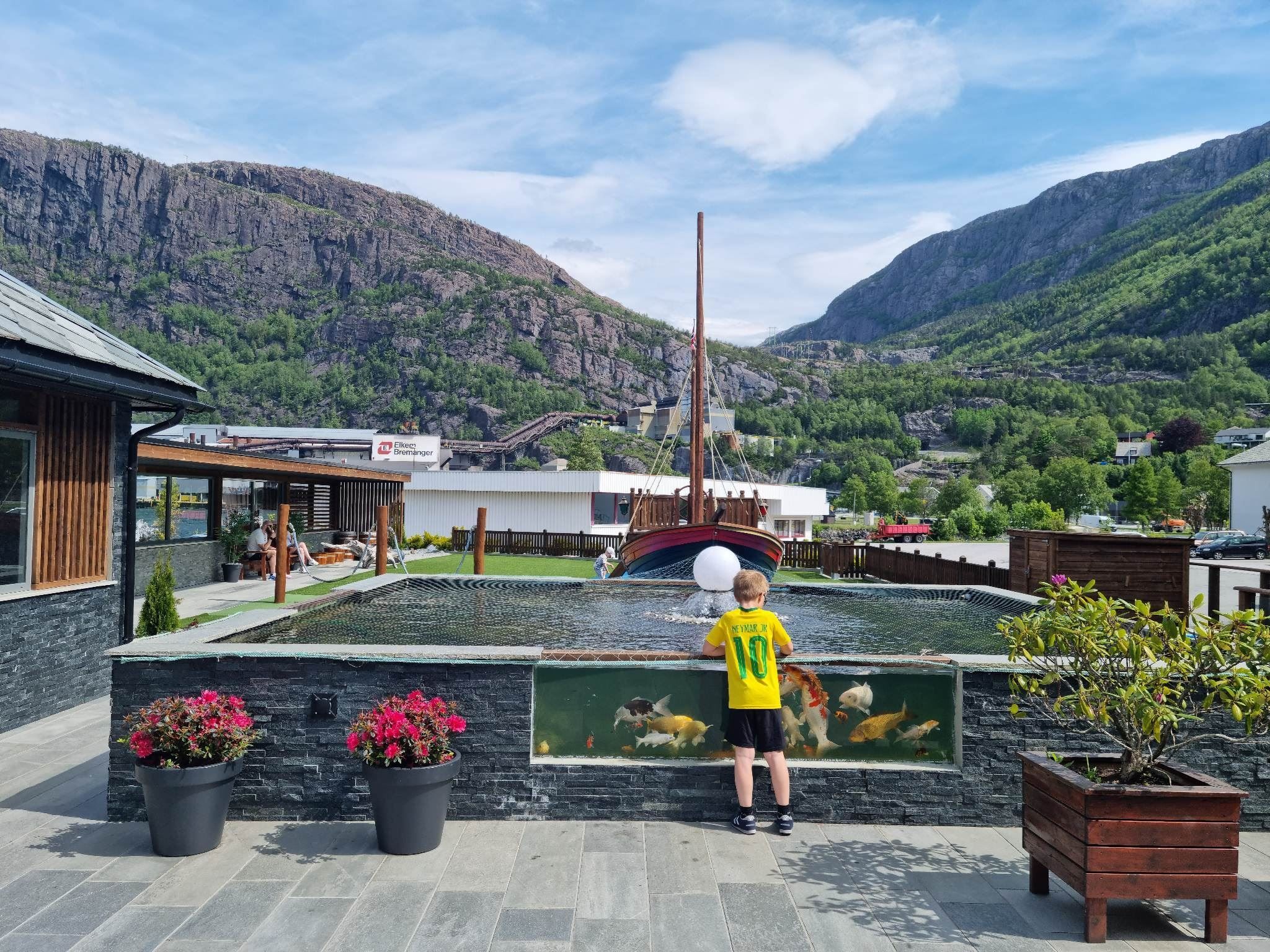 Ein gut i gul trøye står ved ein fiskedam med store fiskar, med fjell, tre og båt i bakgrunnen..