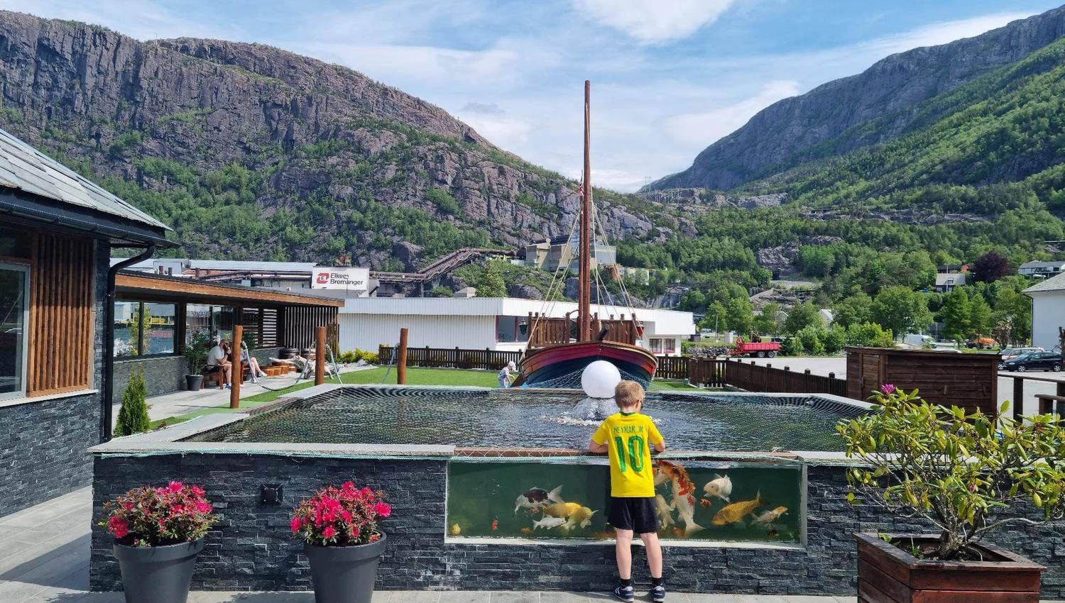 Ein gut i gul trøye står ved ein fiskedam med store fiskar, med fjell, tre og båt i bakgrunnen..