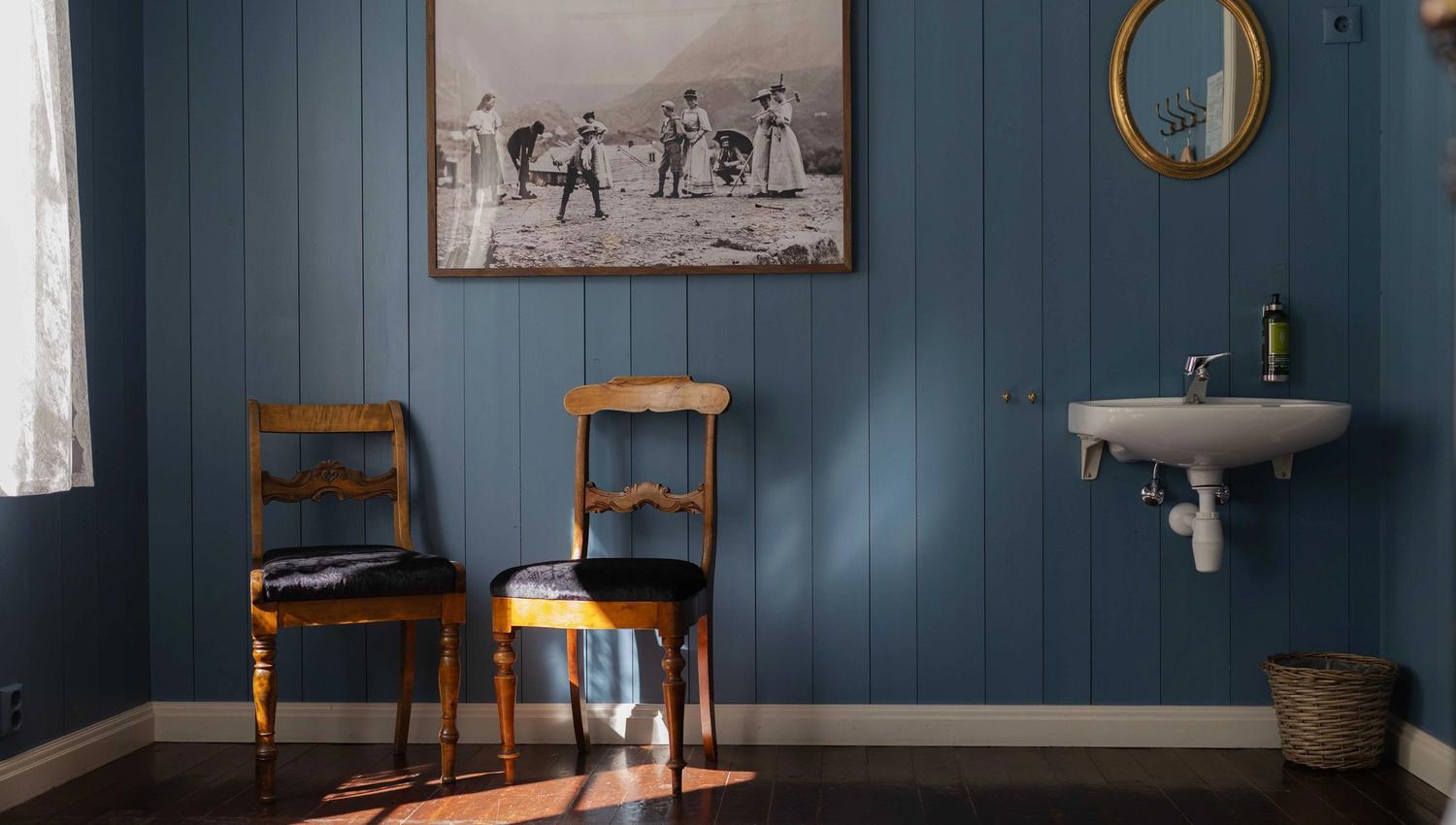 Detaljar frå eit rom i Fruehuset – tidstypiske møblar, veggfaste bilete og varme solstrålar over tregolv.