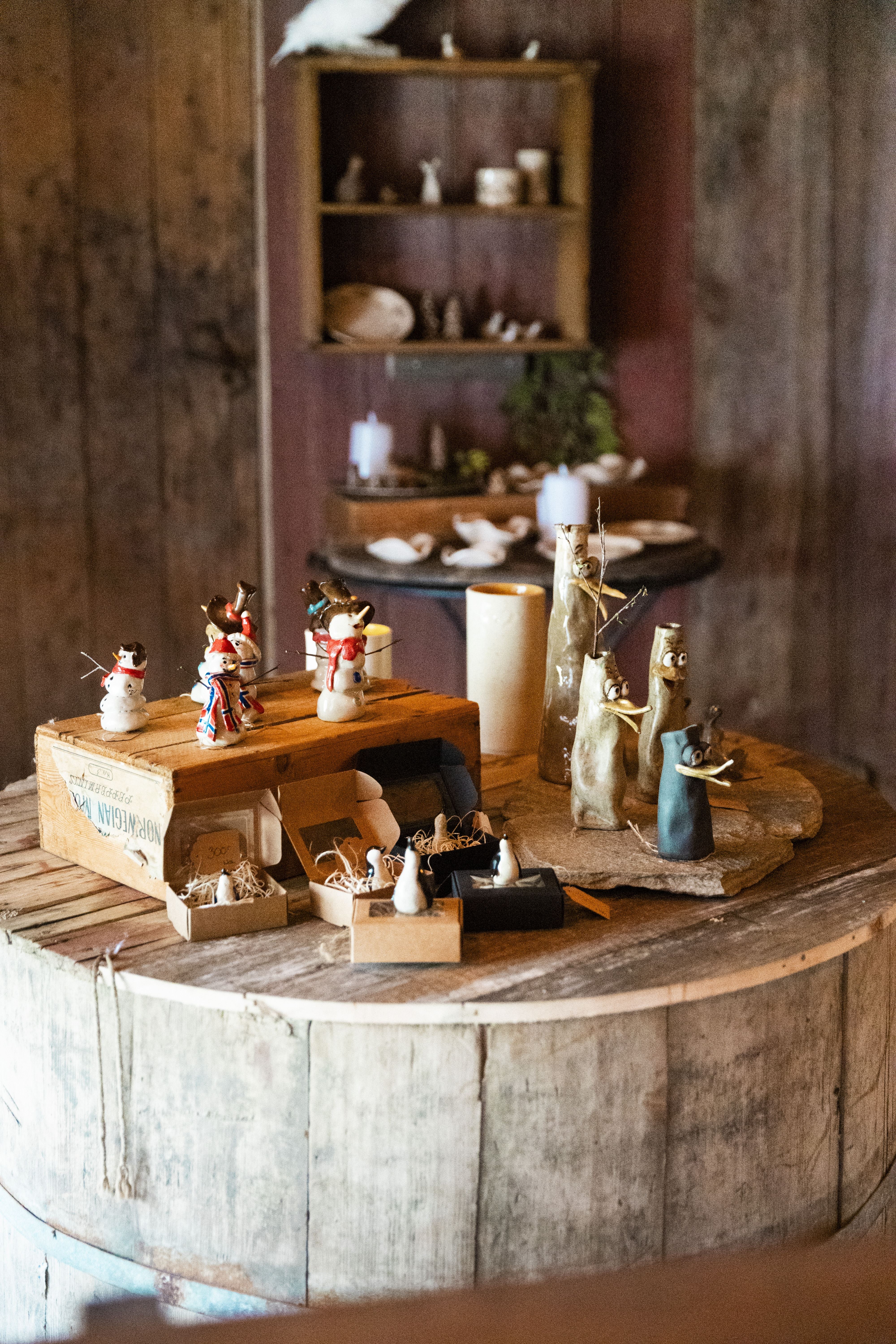 Small handmade figurines and crafts displayed on a round wooden table inside a rustic room with shelves behind.