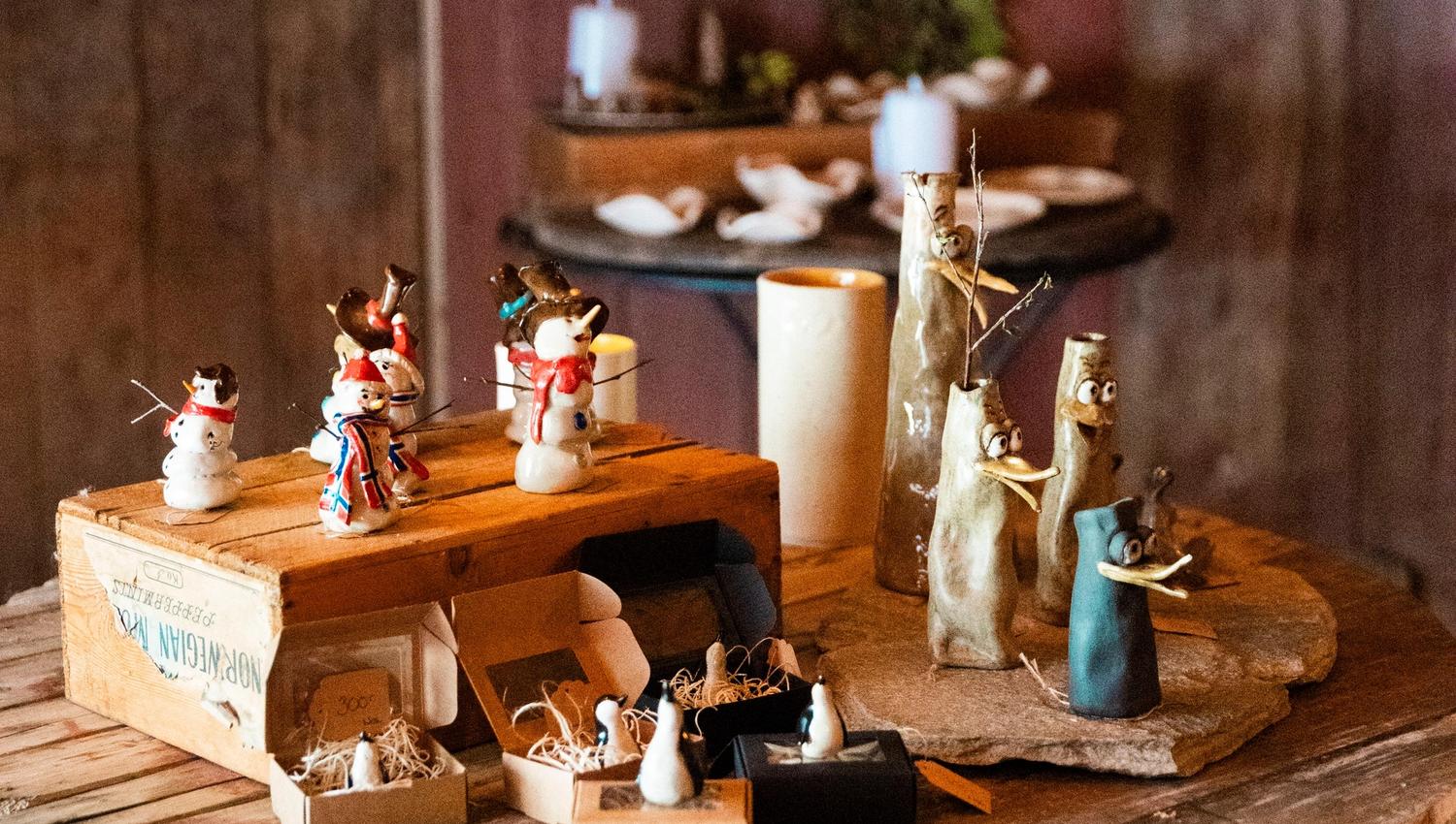 Small handmade figurines and crafts displayed on a round wooden table inside a rustic room with shelves behind.
