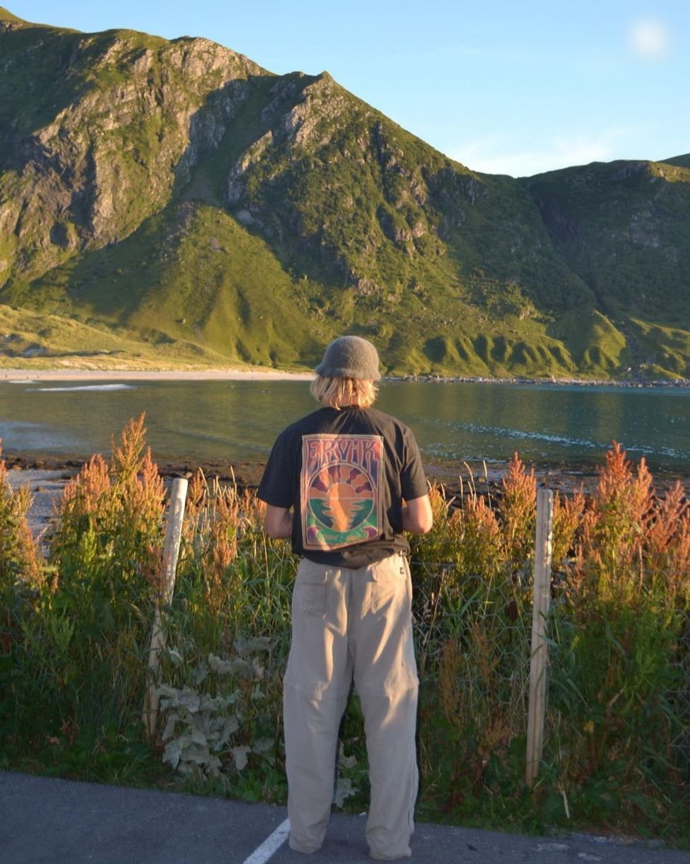 Ein person står med ryggen til ved eit gjerde og ser utover vatn mot grøne fjell i kveldssol ved Ervik Surfshop..