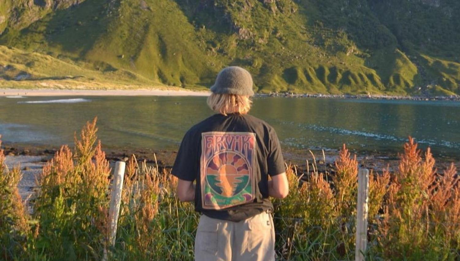 Ein person står med ryggen til ved eit gjerde og ser utover vatn mot grøne fjell i kveldssol ved Ervik Surfshop..