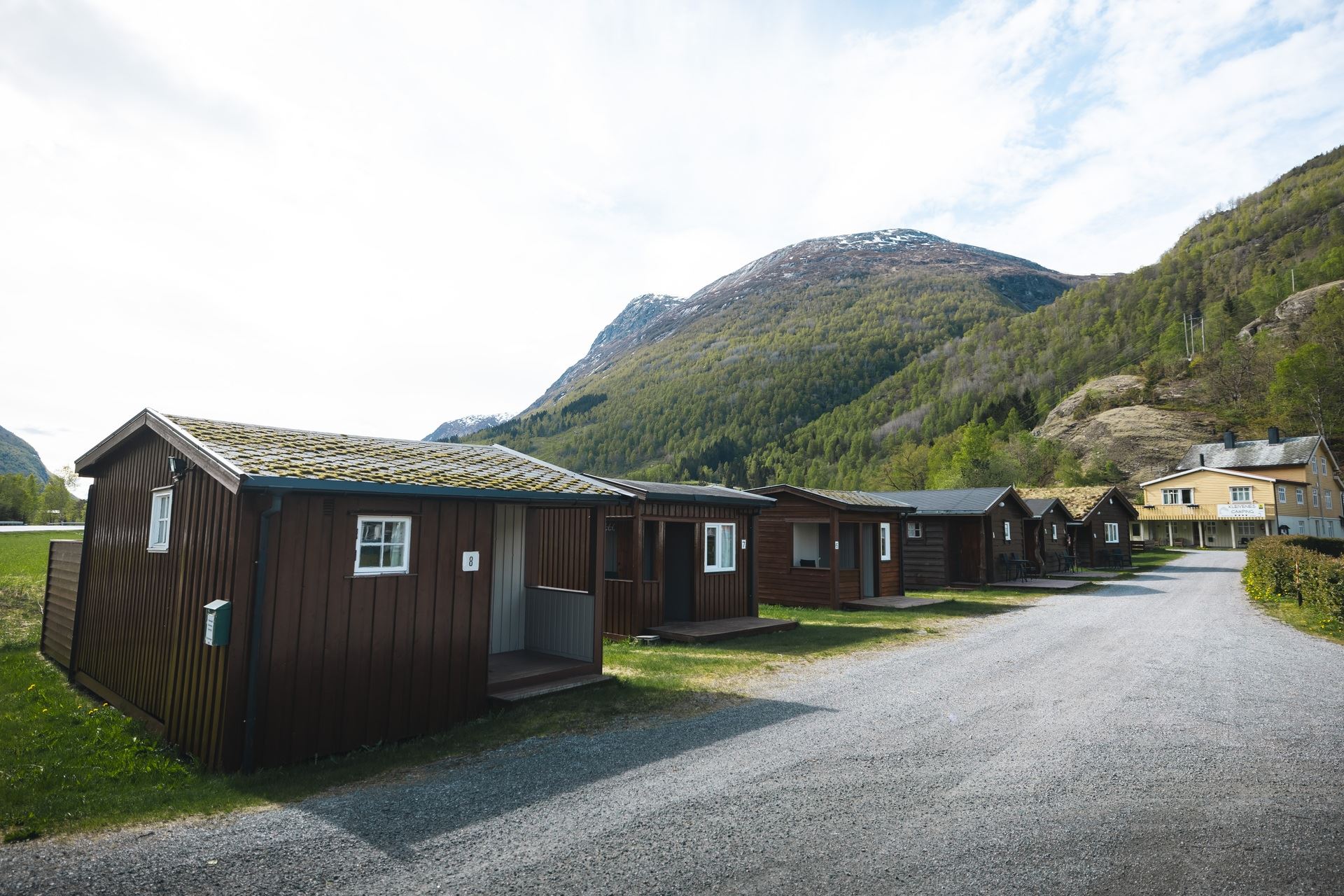 Row of small wooden cabins beside a gravel road, with green forested mountains and a bright sky in the background..