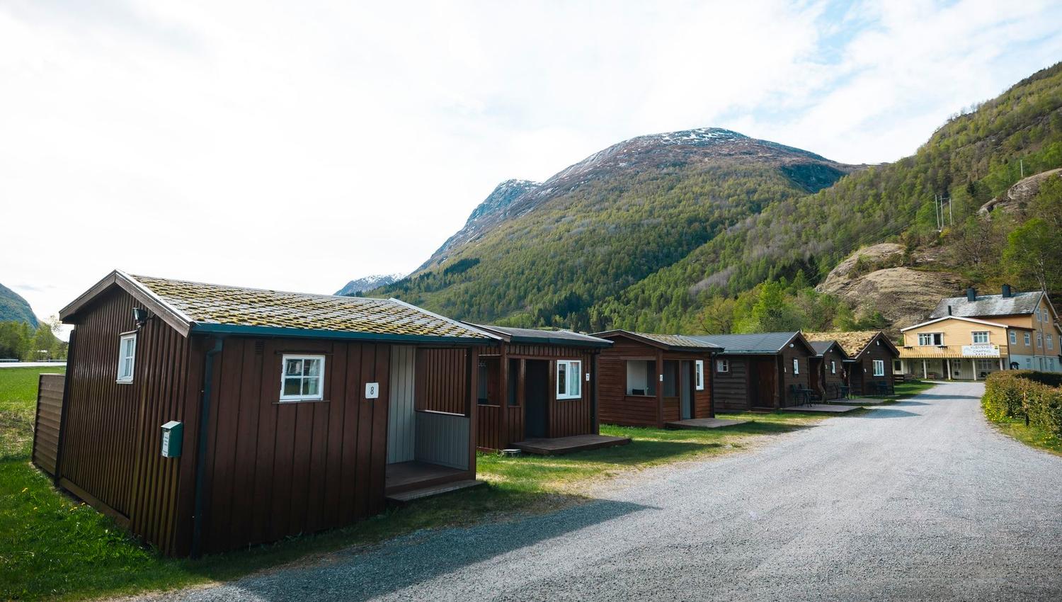 Row of small wooden cabins beside a gravel road, with green forested mountains and a bright sky in the background..