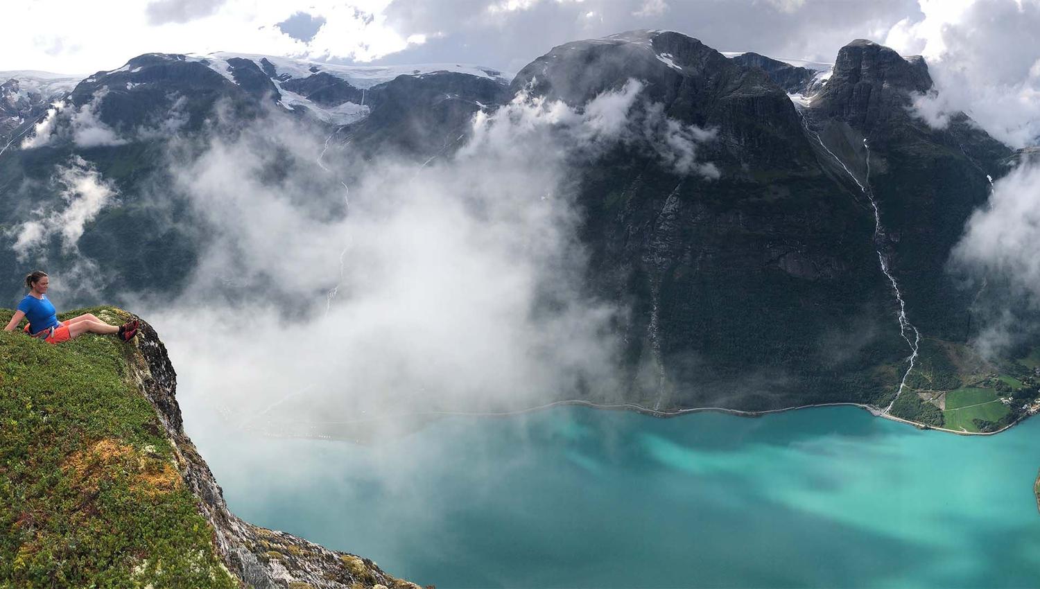 A hiker sits on a grassy cliff overlooking a turquoise fjord with misty clouds and dark mountains in the background..