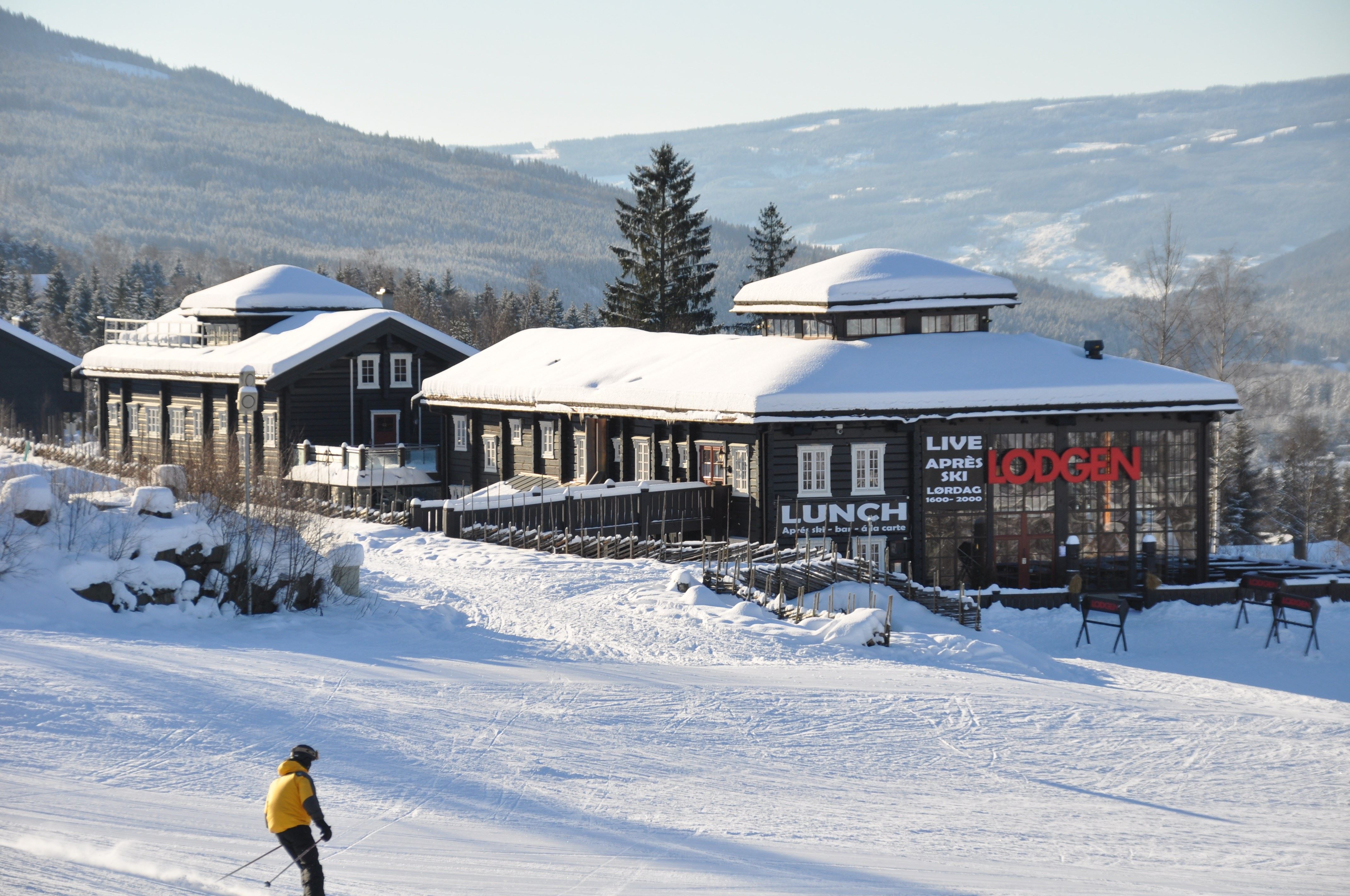 Snötäckt lodge i fjällen med granar och berg i bakgrunden, och en skidåkare i gul jacka i förgrunden.