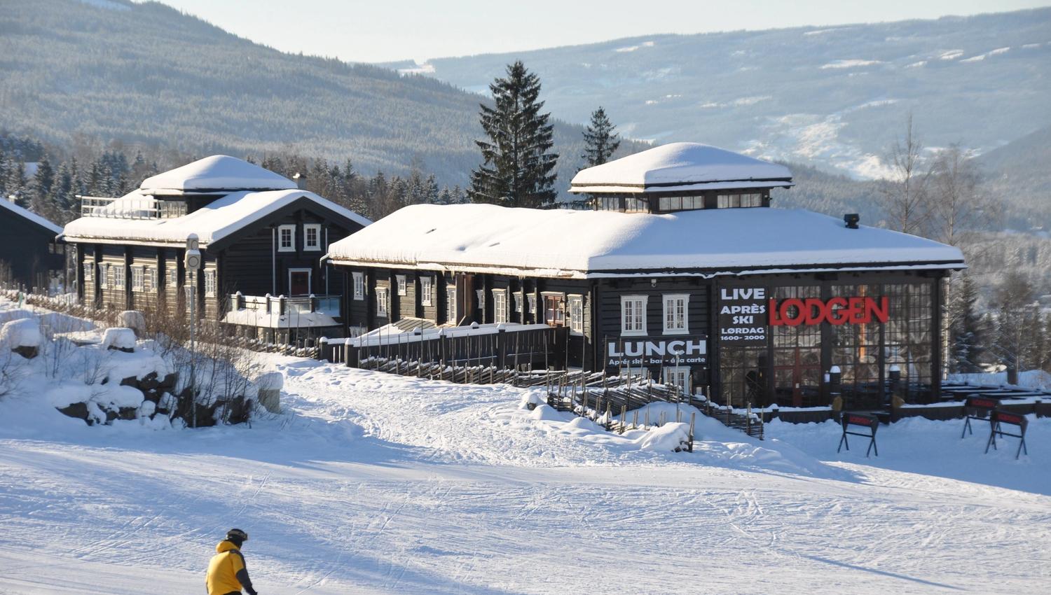 Snötäckt lodge i fjällen med granar och berg i bakgrunden, och en skidåkare i gul jacka i förgrunden.