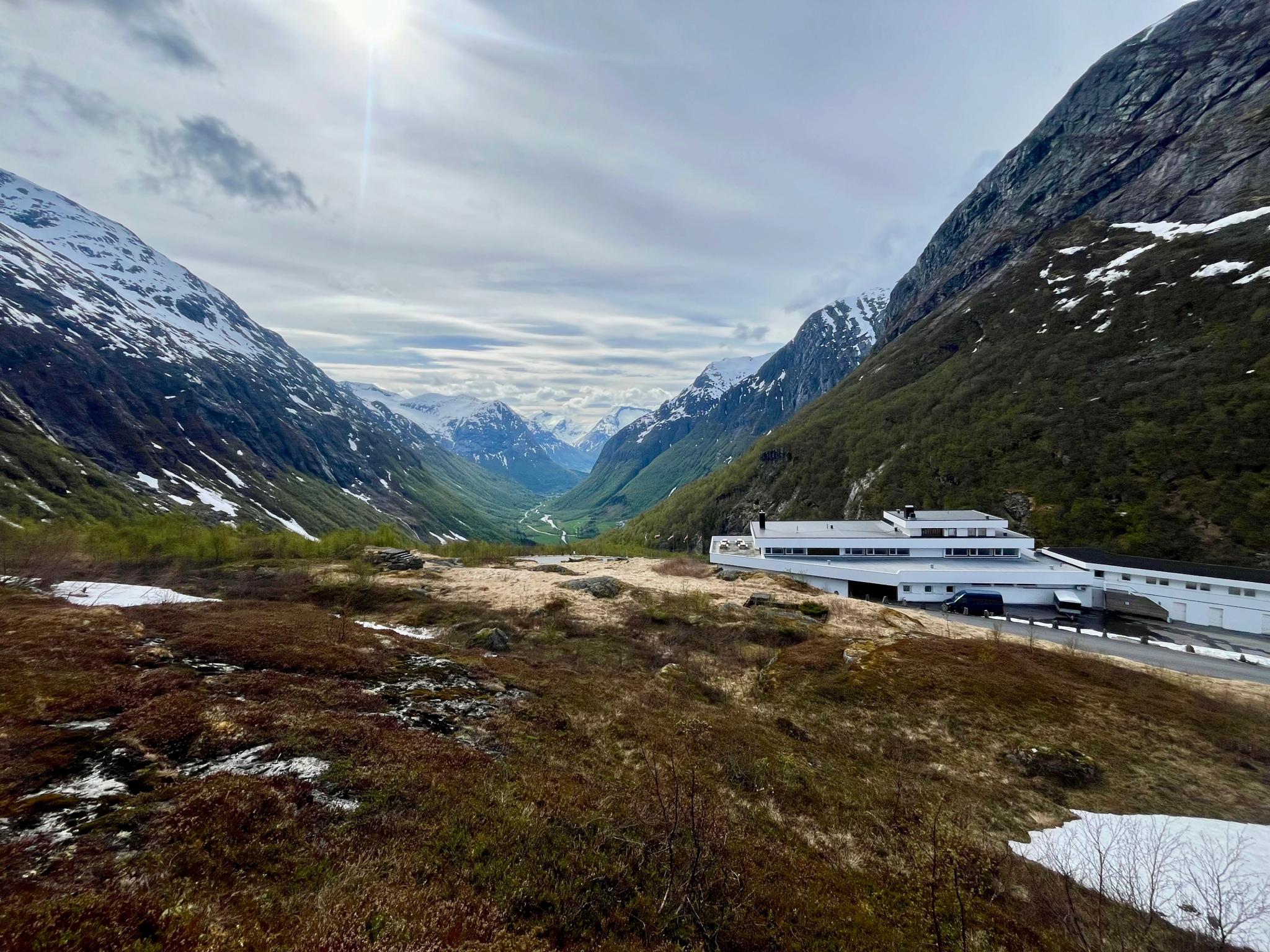 Wide view of a green mountain valley with snow-streaked peaks, cloudy sky, and a white building on a hillside..