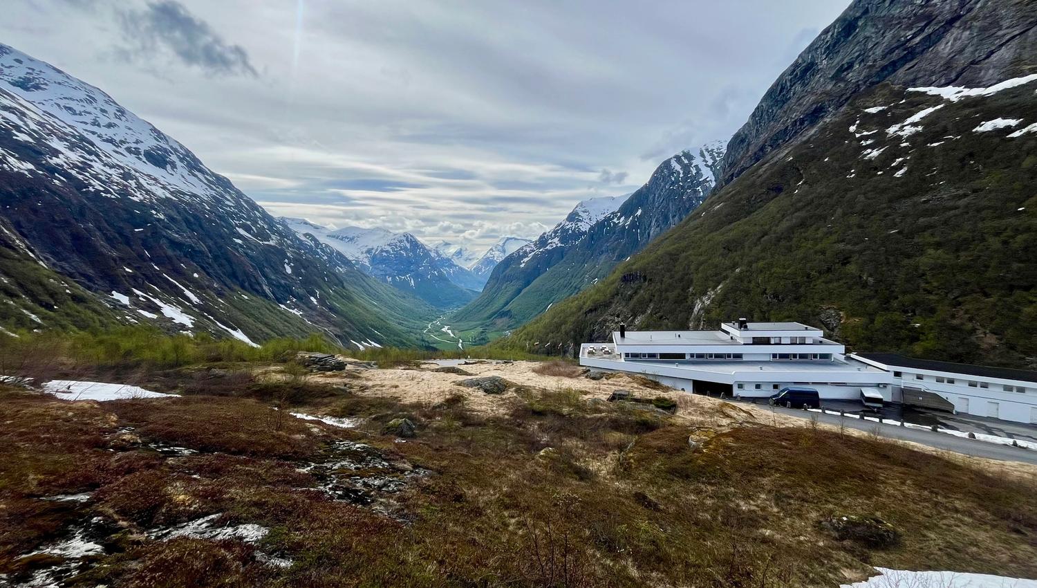 Wide view of a green mountain valley with snow-streaked peaks, cloudy sky, and a white building on a hillside..