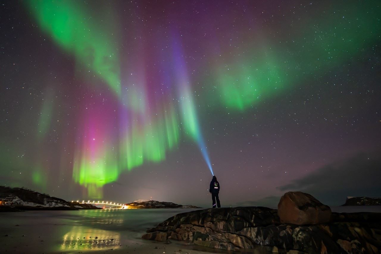 A person wearing a headlamp stands by the water beneath the Northern Lights in the Arctic landscape.