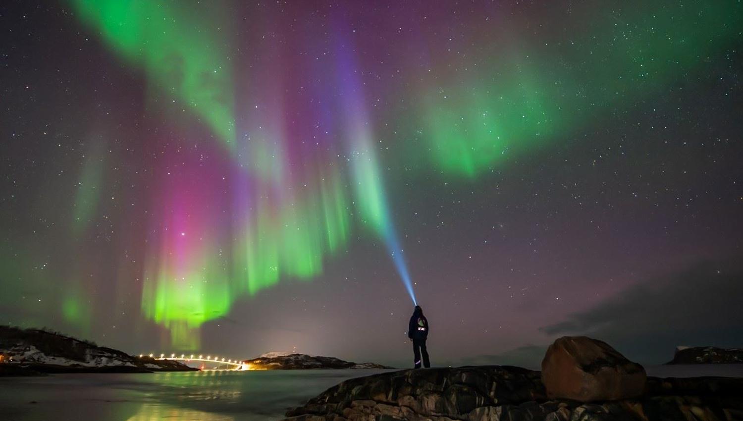 A person wearing a headlamp stands by the water beneath the Northern Lights in the Arctic landscape.