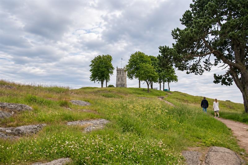 To personer går langs en sti gjennom en blomstrende gresslette mot en hvit kirke på en høyde.