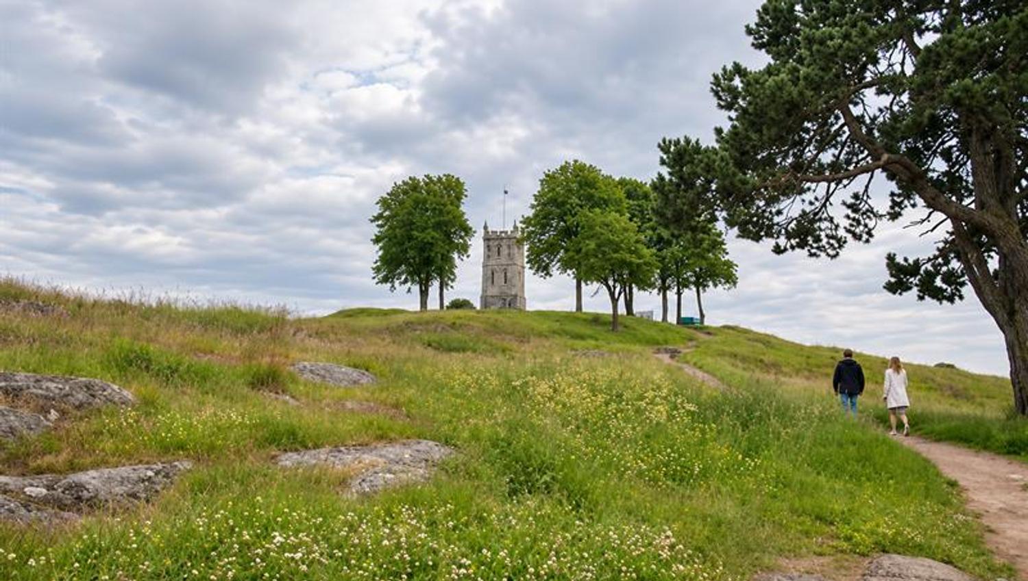 To personer går langs en sti gjennom en blomstrende gresslette mot en hvit kirke på en høyde.
