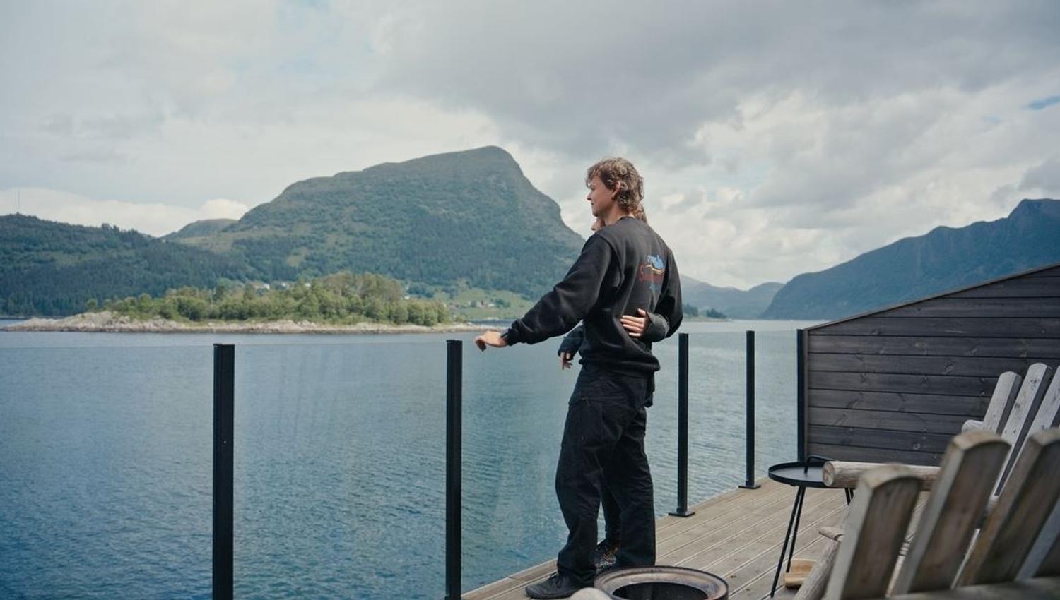 Two people embrace on a wooden deck by calm water, with forested mountains and cloudy sky in the background..