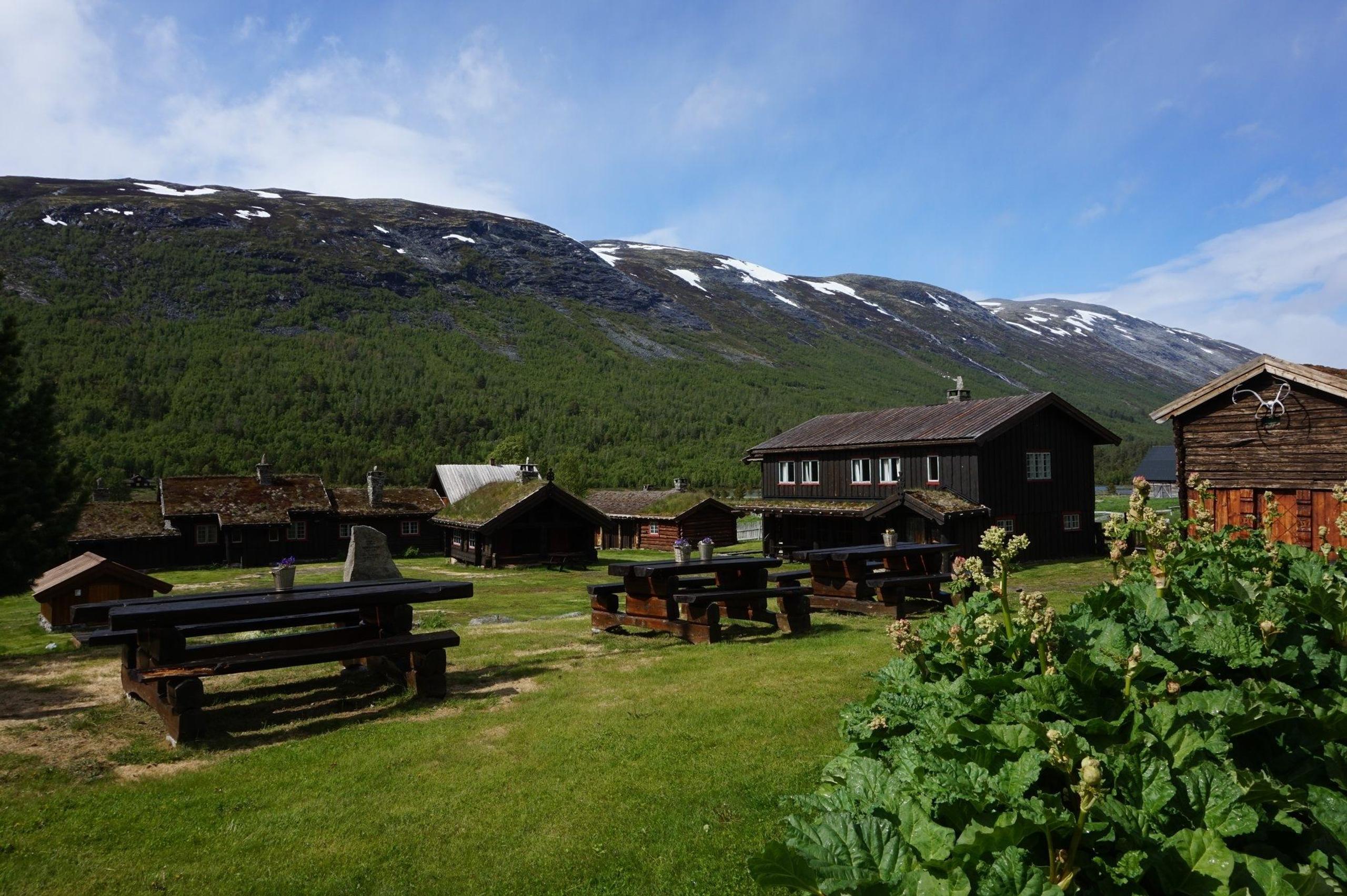 Wooden cabins and picnic tables on green grass, with forested mountains and patchy snow under a blue sky..