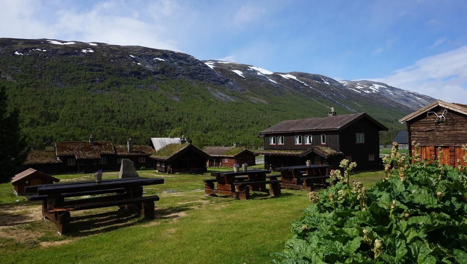 Wooden cabins and picnic tables on green grass, with forested mountains and patchy snow under a blue sky..