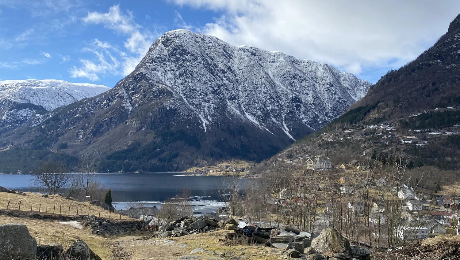 View from the Hjøllo–Mannsåker cultural trail overlooking the Hardanger fjord landscape with snow-covered mountains and blue sky.