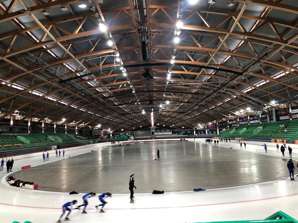 Indoor skating rink with skaters on the ice. Overhead lighting and wooden roof structures are visible above the rink.